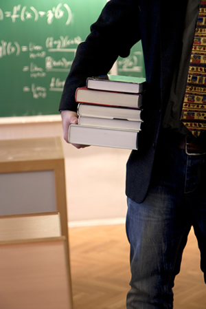 A male holding a stack of books with a chalkboard in the background.
