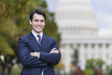 A young politician, staffer, or lobbyist holds with his arms folded, with a confident smile. The United States Capitol building is in the background.
